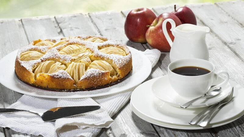 Apfelkuchen, Kaffeetasse und rote Äpfel auf hellem Holztisch