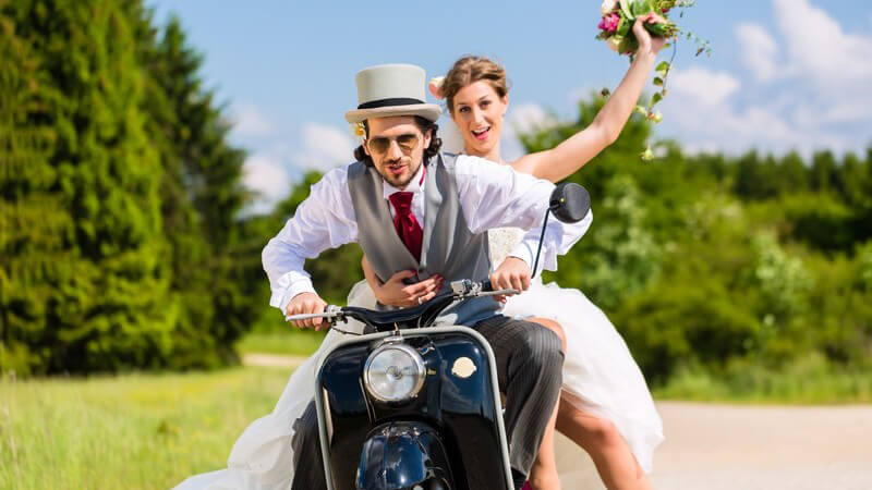 Hochzeitspaar fährt auf einem schwarzen Motorroller durch's Grüne
