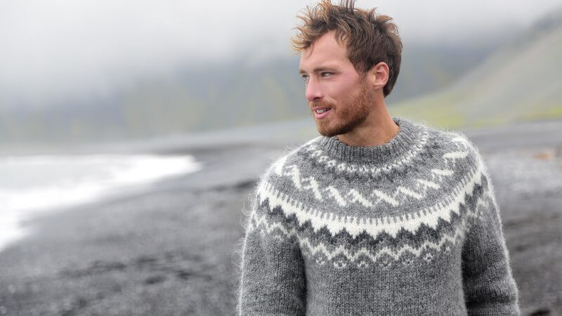 Bärtiger Mann in grauem Wollpulli spaziert an einem schwarzen Strand auf Island