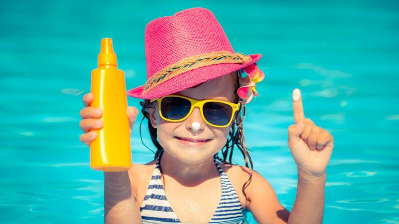 Mädchen mit pinkem Sonnenhut im Pool, hält eine gelbe Sonnencremeflasche hoch
