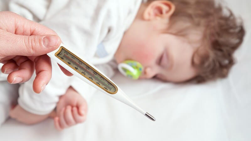 Schlafendes Kleinkind mit grünem Schnuller, im Vordergrund wird ein Fieberthermometer gehalten