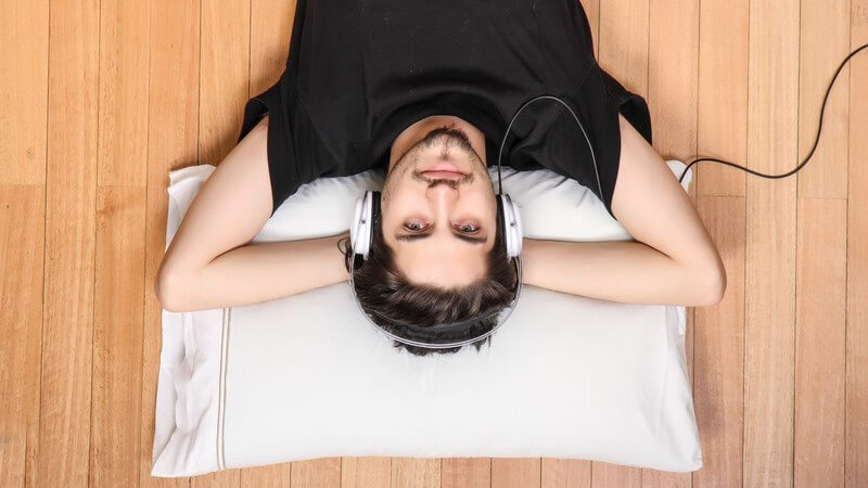 Junger Mann in schwarzem Shirt liegt auf weißem Kissen auf Holzboden und hört Musik über Kopfhörer