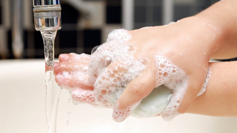 Hände mit Seifenschaum halten Seife, daneben fließendes Wasser