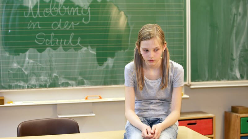 Mobbing in der Schule: Mädchen sitzt traurig und allein auf Tisch in Klassenzimmer
