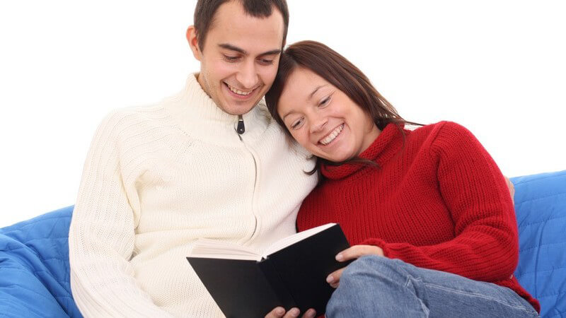 Junges, lächelndes Paar sitzt auf der Couch und liest ein Buch
