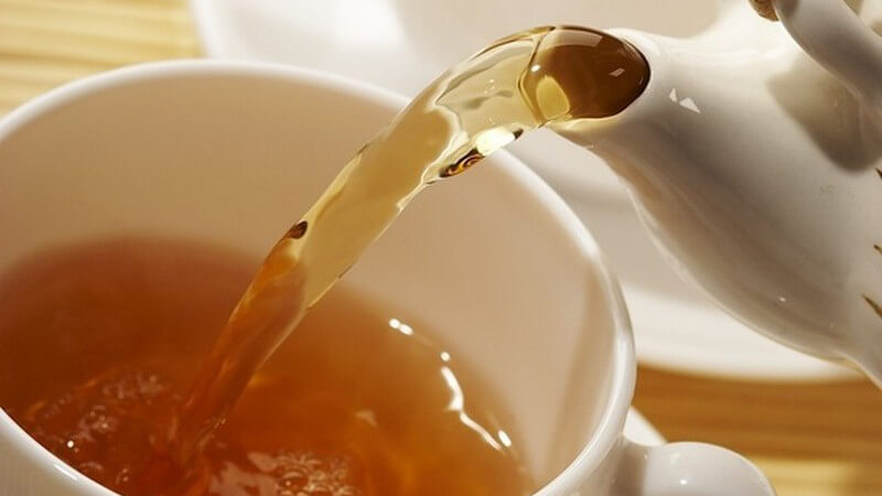 Halbvolle Teetasse vorne, weiße Teekanne schenkt schwarzen Tee o. Early Grey ein, hinten leere Teetasse, auf Holz