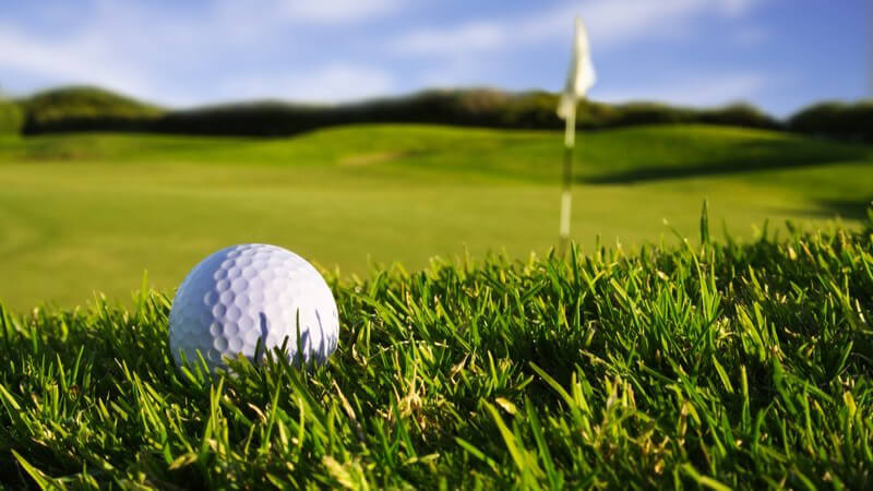 Nahaufnahme Golfball auf grüner Wiese, im Hintergrund Golffahne und blauer Himmel