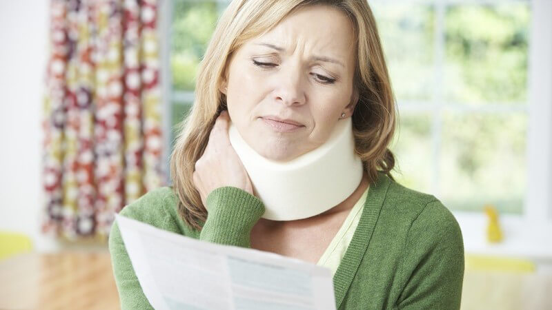 Frau in grüner Strickjacke sitzt mit Schleudertrauma zu Hause, fasst sich an die Halskrause und liest einen Zettel