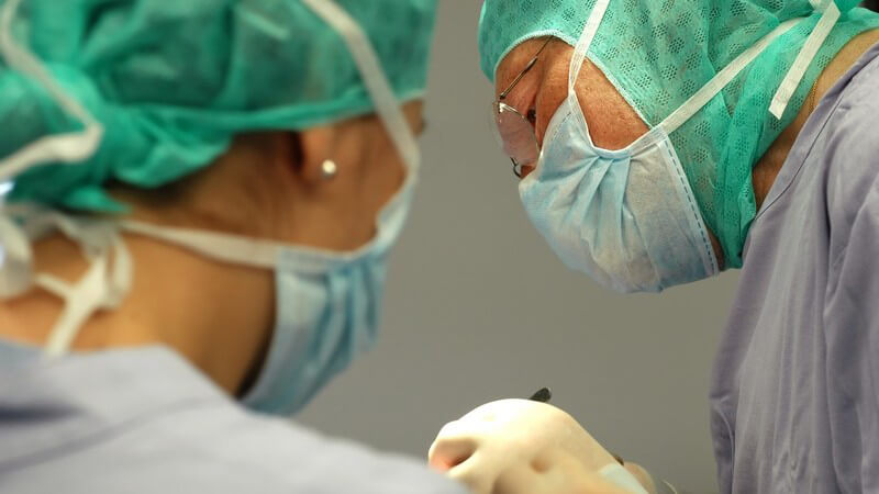 Chirurg im Profil am OP-Tisch, im Vordergrund eine Chirurgin von hinten gesehen