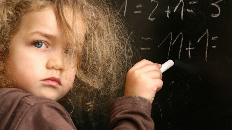 Mädchen mit zerzausten Haaren und ernstem Blick schreibt mit Kreide an die Tafel
