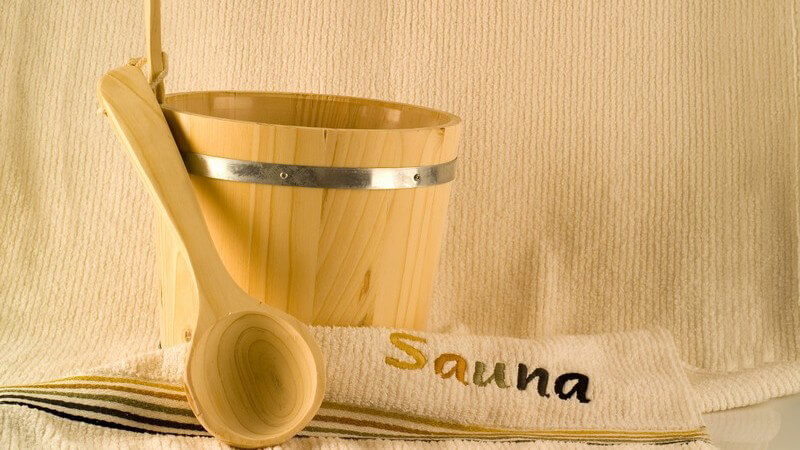 Holzeimer, Holzlöffel und Handtuch mit der Aufschrift "Sauna" vor beigem Hintergrund