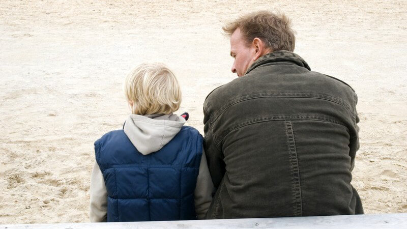 Rückansicht Vater und Sohn sitzen auf Bank vor Sand