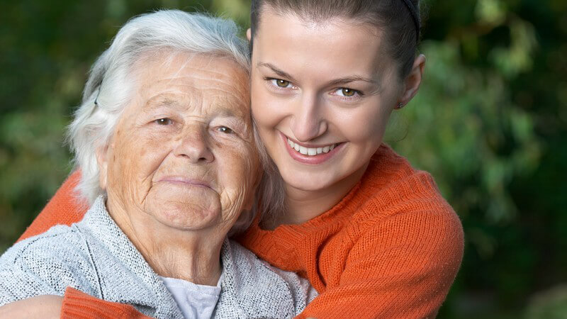 Alte Frau mit weißen Haaren wird von Tocher im Arm gehalten, beide lächeln in die Kamera