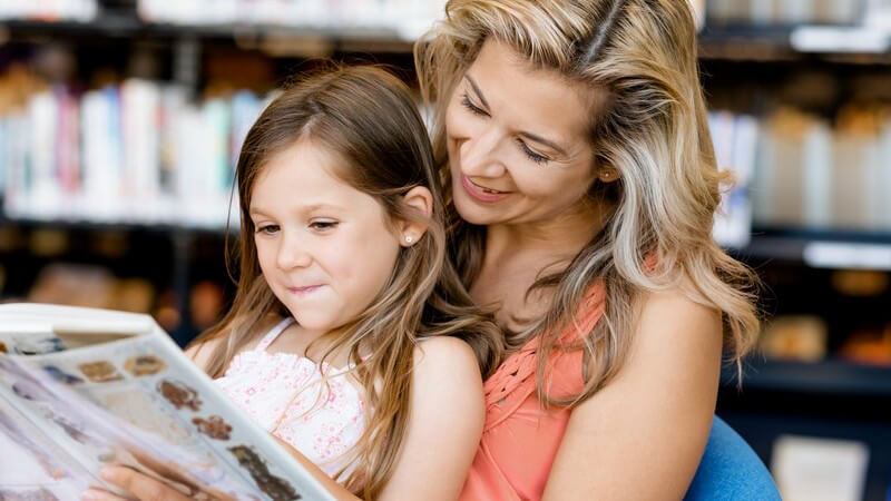 Mutter sitzt mit ihrer Tochter auf dem Schoß in einer Bibliothek und liest mit ihr ein Kinderbuch