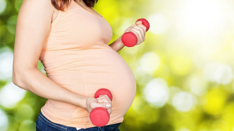 Schwangere Frau mit zwei kleinen roten Kurzhanteln im Grünen