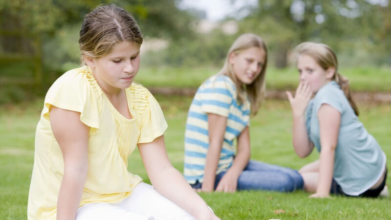 Mädchen sitzt traurig auf Wiese und wird von Mädchen im Hintergrund ausgelacht
