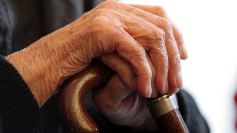 Faltige Hände eines alten Menschen o Seniors mit schwarzer Bekleidung an Gehstock o Krückstock