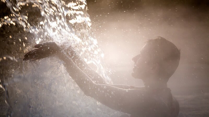 Frau badet unter einem Wasserfall im Thermalbad, verdeckt von Gischt und Nebel