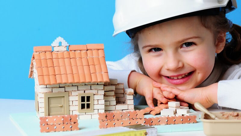 Kleines Mädchen mit Bauarbeiter Helm neben Spielzeughaus