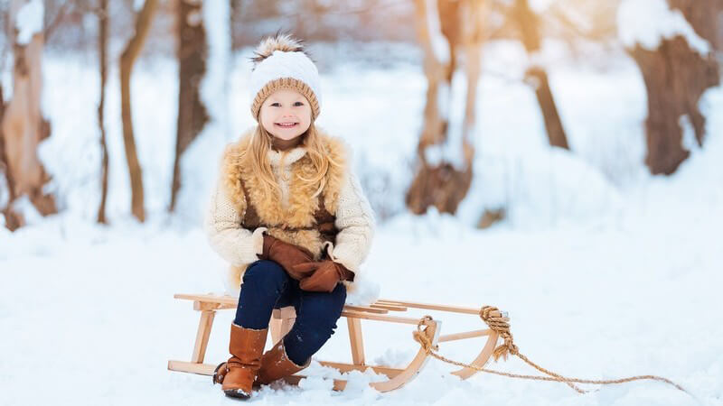 Kleines Mädchen in dicker Winterkleidung posiert lächelnd auf einem Holzschlitten im Schnee