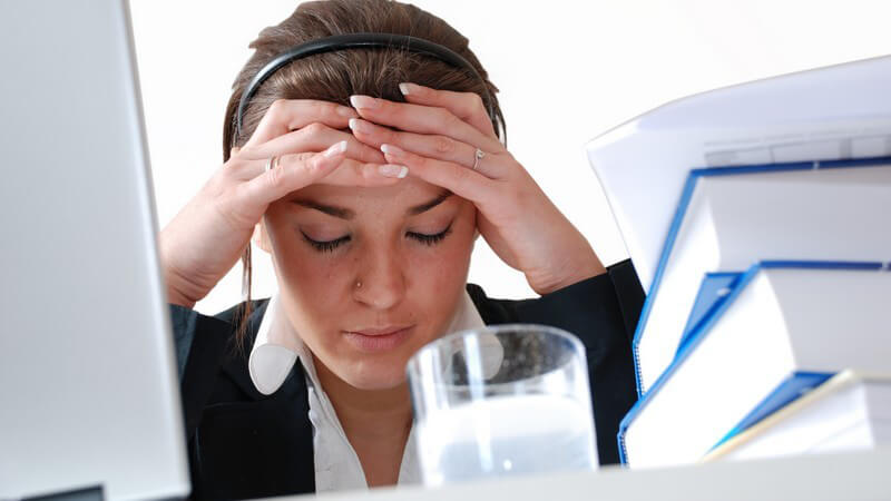 Junge dunkelhaarige Frau mit Bluse in Büro zwischen Computer und Büchern mit Wasserglas, hält sich Kopf wg Kopfschmerzen