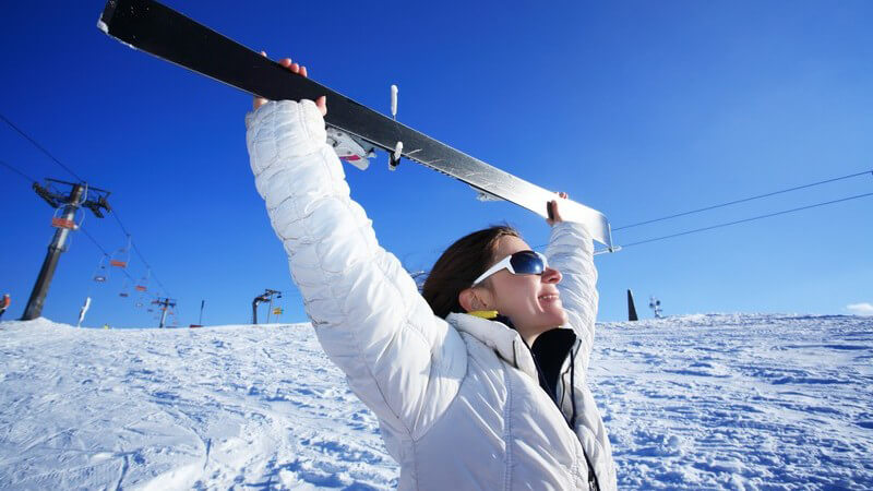 Junge Frau auf Skipiste hält ihren Skier nach oben in den blauen Himmel