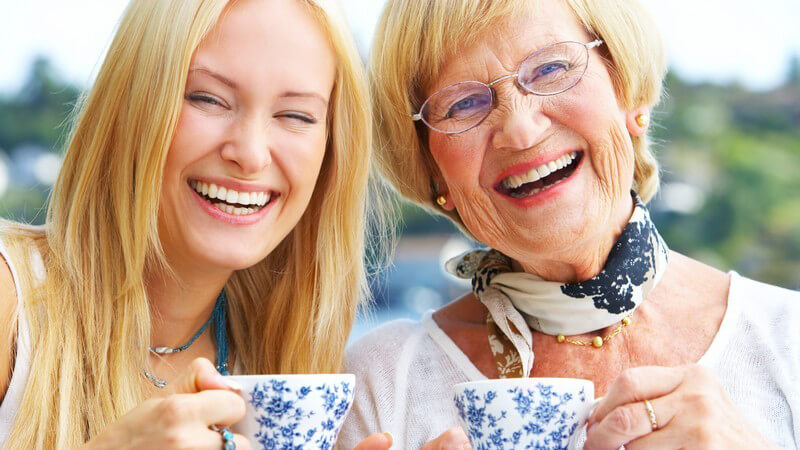 Junge Frau mit Großmutter, beide halten Teetassen in Hand und lachen in Kamera