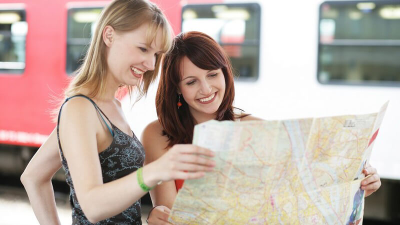 Zwei junge Frauen schauen am Bahnhof auf Stadtplan, im Hintergrund Zug