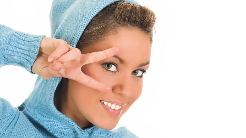 Junge Frau in blauem Kapuzenshirt hält zwei Finger an rechtes Auge
