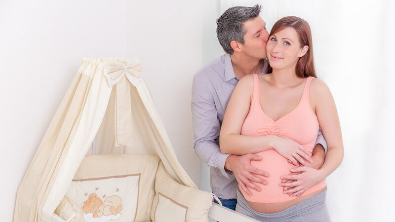 Werdender Vater umklammert und küsst seine schwangere Frau neben dem leeren Babybett