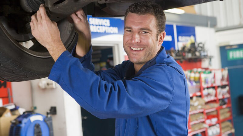 Automechaniker im Blaumann an Hebebühne unter Auto, lächelnd