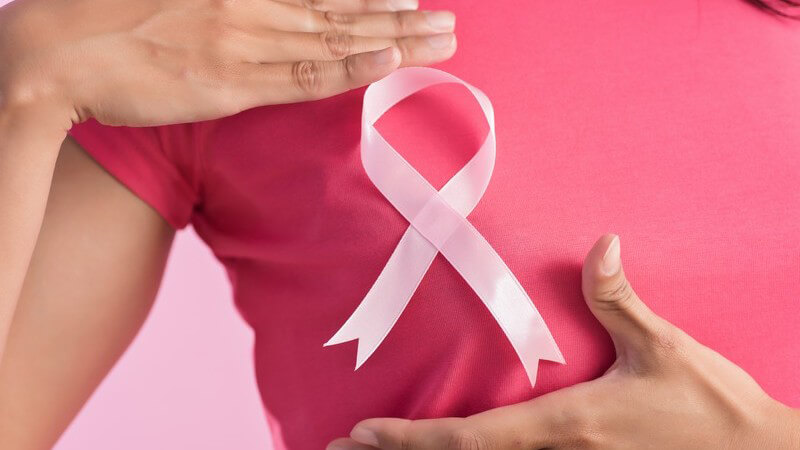 Frau in pinkem Shirt trägt eine rosa Brustkrebs-Schleife auf der Brust und hält die Hände daneben