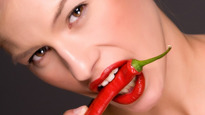 Blonde Frau mit roter Chili Schote im Mund mit rotem Lippenstift