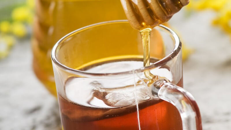 Glas mit Tee, Honig wird von Honiglöffel hineingegossen, im Hintergrund Glas mit Honig