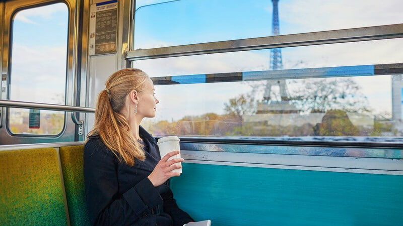 Blonde Frau sitzt mit Coffee-to-go-Becher in einer Pariser Straßenbahn und guckt aus dem Fenster