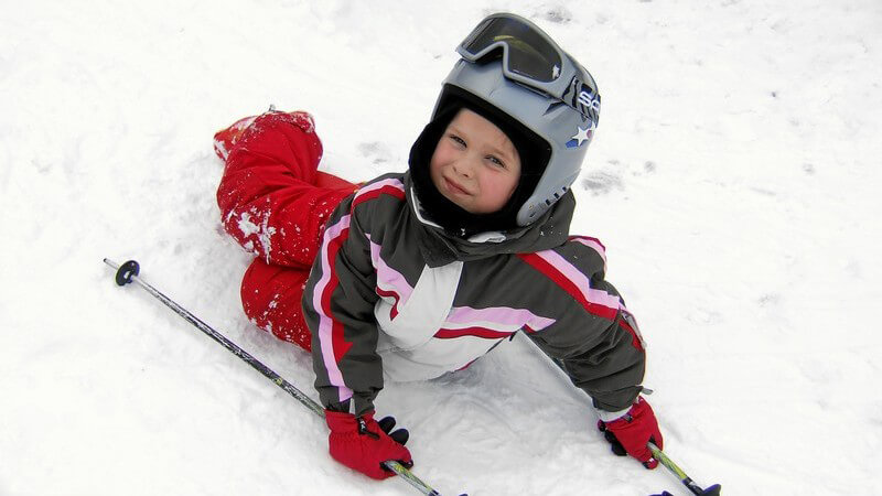 Kleiner Junge liegt im Schnee, Skihelm, Skistöcke, Skibrille