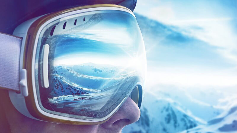 Seitliche Nahaufnahme einer Skibrille auf dem Gesicht eines Mannes, in der Brille spiegeln sich Sonne und Skigebiet