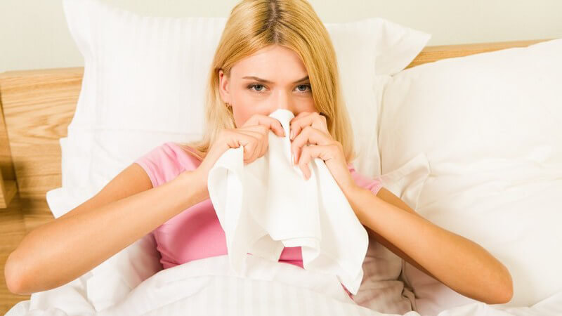 Junge blonde Frau liegt krank im Bett und putzt sich die Nase