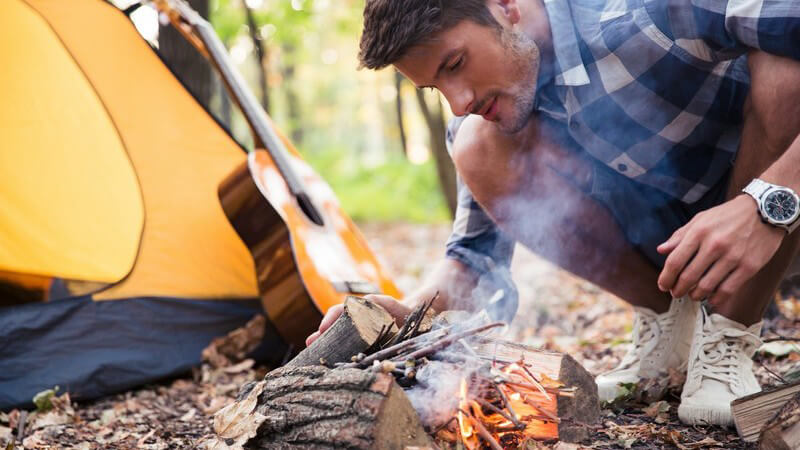 Junger Mann in Karohemd entzündet ein kleines Lagerfeuer neben einem Zelt im Wald