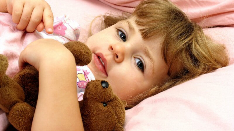 Kleines Mädchen liegt mit Teddybär im Arm im Bett und schaut in Kamera