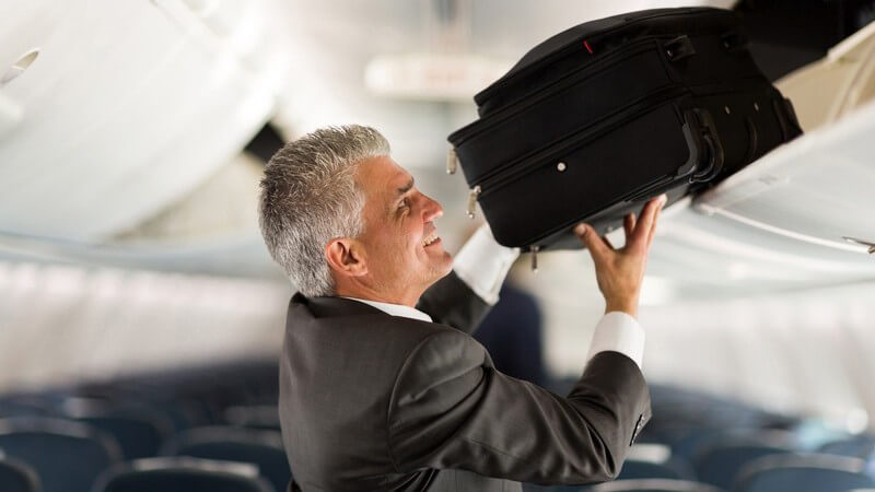Älterer Geschäftsmann verstaut seinen kleinen Koffer im Handgepäckfach eines Flugzeugs