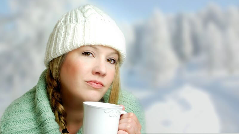 Junge Frau mit Zöpfen und weißer Strickmütze hält Becher Tee in Hand, im Hintergrund Bergpanorama