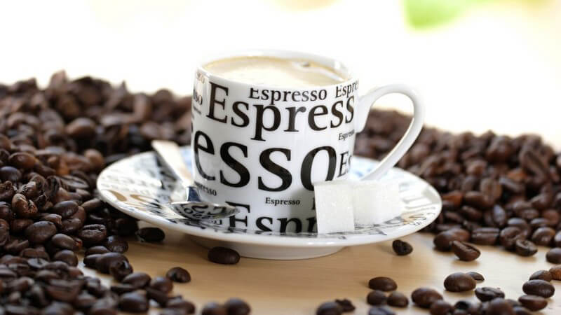 Espresso in Tasse mit Löffel, Zucker auf Tisch, umgeben von Kaffeebohnen