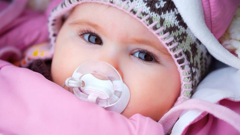 Baby in Winterkleidung und Schnuller im Mund schaut in Kamera