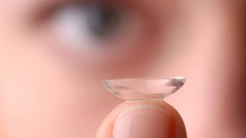 Kontaktlinse auf Fingerspitze im Vordergrund, Auge unscharf im Hintergrund
