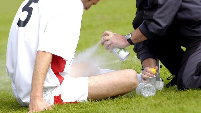 Fußballspieler sitzt am Boden und ist verletzt, sein Bein wird mit Kühlspray gekühlt