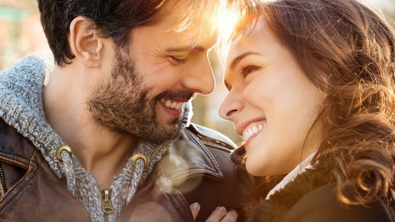 Glückliches Paar in der Herbstsonne sieht sich verliebt an, er mit brauner Lederjacke über grauer Kapuzenstrickjacke