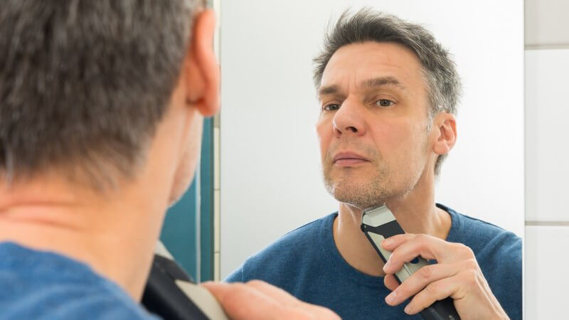 Reifer Mann in blauem Pullover steht vor dem Spiegel und trimmt sich den Bart mit einem Rasierapparat