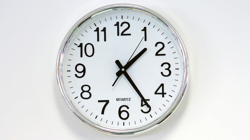 Weiße Wanduhr mit schwarzer Aufschrift, zeigt Uhrzeit fünf vor halb zwei