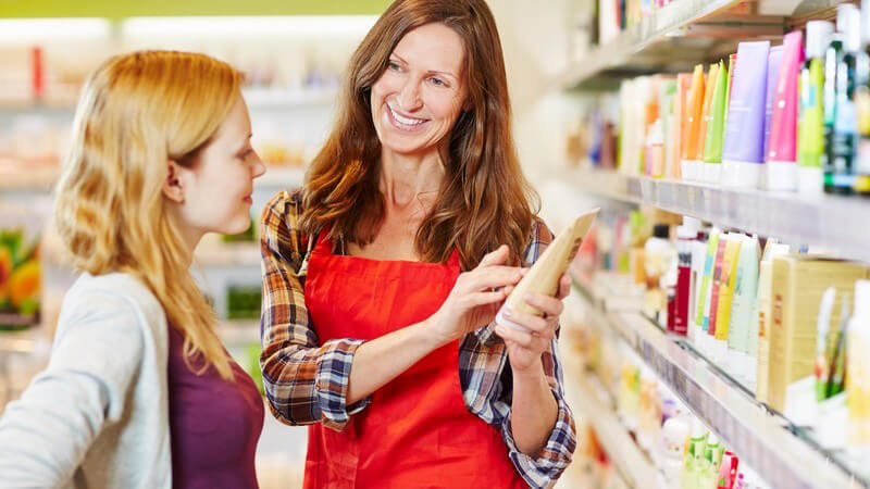 Verkäuferin mit roter Schürze berät eine junge Frau an einem Regal im Drogeriemarkt